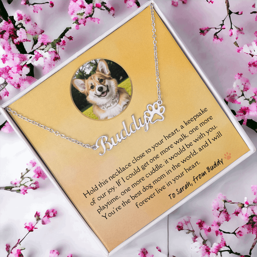 Silver custom dog memorial necklace with engraved name and paw print charm – pet loss keepsake with a golden colored message card with a heartfelt message from dog to his dog mom. Pink flowers in the background.